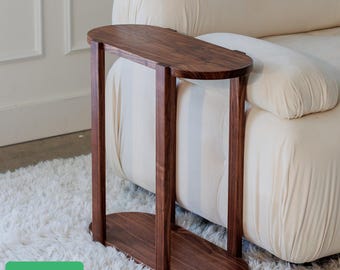 May include: A dark brown, oval-shaped side table with a minimalist design. The table has a rectangular base and top, connected by two vertical supports. The table is made of wood and is placed next to a cream-colored sofa.