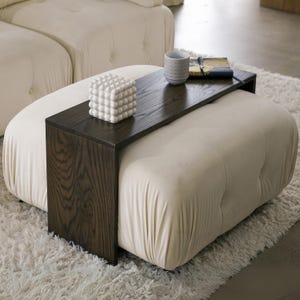 Puede incluir: Un otomano blanco con una bandeja de madera oscura unida a la parte superior. La bandeja tiene un cubo decorativo blanco y un libro encima.