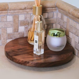 May include: A round, dark brown wooden tray holds a clear glass diffuser bottle with reeds, a perfume bottle, and a frosted glass jar with a gold lid. The items are arranged on a light-colored surface against a tiled wall.