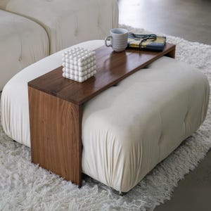 May include: A white ottoman with a tufted top and a wooden tray attached to the side. The tray has a white decorative cube and a gray mug on it.