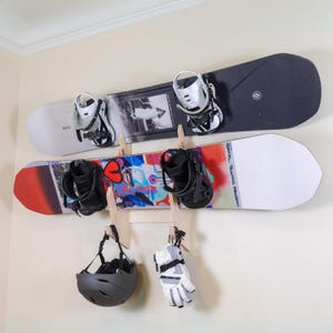 May include: Two snowboards with bindings are mounted on a wall, above a black helmet and a pair of gloves. The top snowboard is black and white, and the bottom one is red, white, and blue. The helmet and gloves are hanging below.