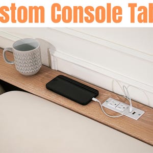 May include: A custom console table with a light wood finish, featuring a built-in power outlet and USB charging ports. A gray mug and a black smartphone are on the table. The text "Custom Console Table" is at the top.