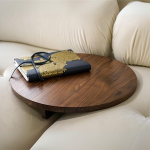 May include: A round wooden side table with a dark brown finish. The table is attached to a white couch and has a book on top.