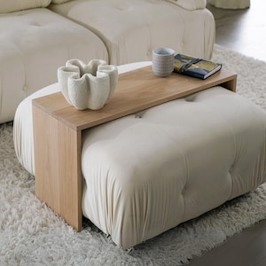 May include: A white upholstered ottoman with a wooden tray attached to the top. The tray has a white ceramic bowl and a small book on it.