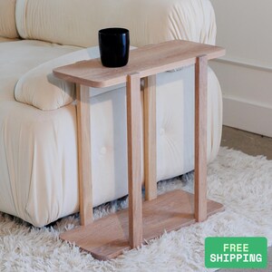 Puede incluir: Una mesa auxiliar de madera clara con una parte superior rectangular y un estante inferior. La mesa tiene un diseño minimalista con cuatro soportes verticales. Un vaso negro está sobre la mesa. La mesa está sobre una alfombra blanca.