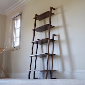 May include: A dark brown wooden leaning shelf with four shelves. The shelves are rectangular and supported by a ladder-like frame. The shelf is leaning against a light-colored wall in a room with a window.