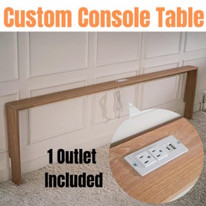 Peut inclure: Une table console personnalisée en bois avec une prise électrique intégrée. La table est fixée au mur et a une finition en bois clair. Le texte "1 Outlet Included" est visible sur l'image.