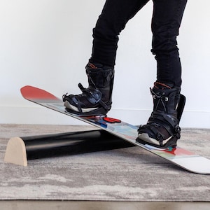 May include: A snowboard with black boots attached, balanced on a black roller with a wooden end. The snowboard has a red and white design on the tip and a colorful graphic in the center. The person is wearing black pants.