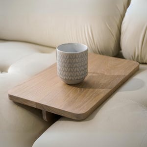 May include: A light brown wooden tray with a handle rests on a cream-colored sofa. A gray and white patterned mug sits on the tray. The mug has a repeating leaf design.