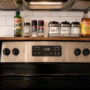 Custom walnut kitchen storage shelf designed to sit above the stove back panel and provide space for spices oils vinegar jars and small decor a practical wooden organizer for a cleaner more organized cooking area