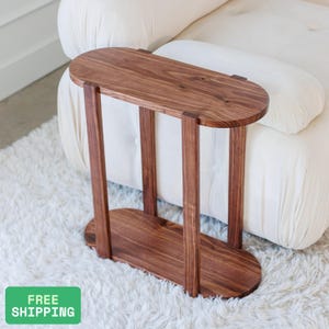 May include: A wooden side table with a dark brown finish. The table has an oval top and base, connected by four vertical supports. The table is designed to fit over the arm of a sofa. The table is made of wood.