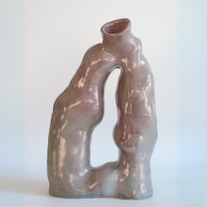 May include: A unique, abstract ceramic vase in shades of mauve and gray. The vase has an organic, flowing shape with a large opening in the center and a tilted top. The glossy finish reflects light, highlighting the curves and textures of the handmade pottery.