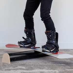 Puede incluir: Una persona con botas de snowboard negras y pantalones negros está de pie sobre una tabla de snowboard. La tabla de snowboard es blanca con detalles en rojo, azul y verde. Debajo hay una tabla de equilibrio negra con un extremo de madera.