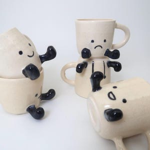 May include: A collection of whimsical ceramic mugs with cartoon-like faces and black accents. The mugs are off-white with black arms and legs, and some have smiling or sad expressions. The mugs are stacked and arranged on a white surface.