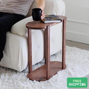 May include: A dark brown, wooden side table with an oval top and base, connected by two vertical supports. A black mug and a notebook rest on the table's surface. The table is positioned next to a white sofa on a fluffy white rug.