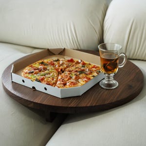 Puede incluir: Una mesa redonda de madera oscura con una pizza en una caja de cartón y un vaso de té. La pizza está cortada en rodajas y tiene ingredientes. La mesa está sobre un sofá blanco.