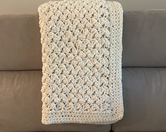 May include: A white crocheted throw blanket with a textured pattern, folded and placed on a light brown couch.