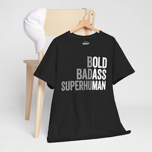Op de afbeelding: Zwart T-shirt met de woorden "BOLD BADASS SUPERHUMAN" in een gelaagd, grijstinten lettertype. Het shirt hangt aan een houten kleerhanger. Een witte baseballpet rust op de rug van een houten stoel.