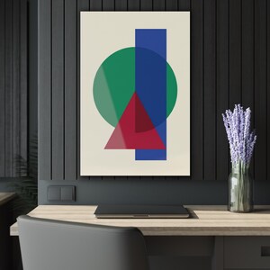 May include: Abstract geometric art print featuring a green circle, a red triangle, and a blue rectangle overlapping on a cream background. The artwork is displayed on a wall above a desk with a laptop and a vase of lavender.