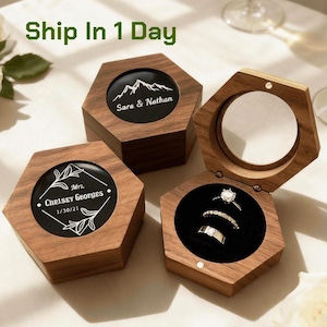 May include: Three hexagonal wooden ring boxes with dark wood grain. Two boxes are closed with custom white engravings: one shows a mountain range with the text "Sara & Nathan," the other a floral frame with "Mrs. Chelsey Georges 1/30/21." One box is open, revealing three silver rings.