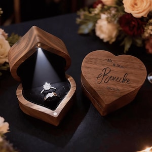 May include: A heart-shaped wooden ring box with a built-in light illuminating two diamond rings. The box is open, revealing a black velvet interior. A second heart-shaped box is closed, with the inscription "Mr. & Mrs. Benecke 09.26.26".