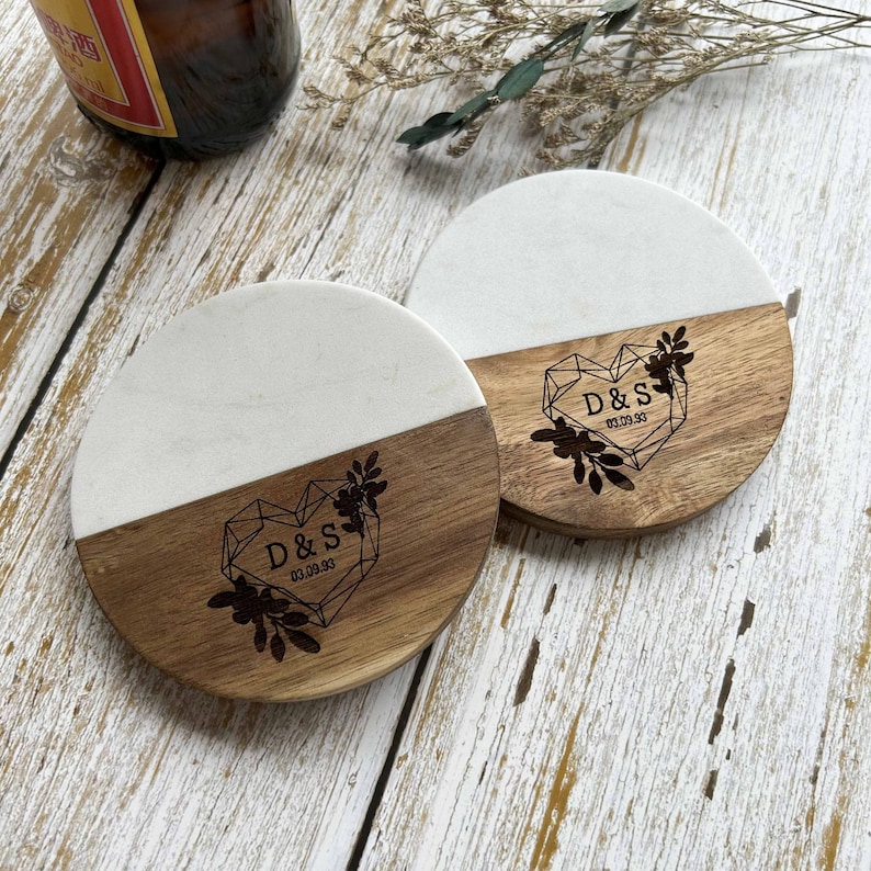 Pu&ograve; includere: Due sottobicchieri rotondi con un top in marmo bianco e un fondo in legno. Il fondo in legno presenta un design a forma di cuore con le iniziali "D & S" e la data "03.09.93" incise.