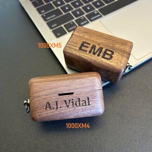 Puede incluir: Dos estuches de madera para auriculares inal&aacute;mbricos, uno grabado con "EMB" y el otro con "A.J. Vidal". Los estuches son de madera marr&oacute;n oscura y tienen un acabado natural. Los estuches est&aacute;n sobre un port&aacute;til plateado.