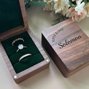 Könnte beinhalten: Eine Holz-Ringbox mit dunkelgrünem Samtinterieur, die drei Ringe enthält. Die Box ist geöffnet und zeigt einen Diamantring und zwei weitere Ringe. Der Deckel der Box ist mit den Namen "Kippa & Jade", "Solomon" und dem Datum "26.09.2025" graviert.