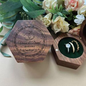 May include: Hexagonal wooden ring box with engraved wedding rings and the names "Vivian & David" and the date "02 OCT 2025". The box is open, revealing two rings on a green velvet interior. A floral arrangement is in the background.