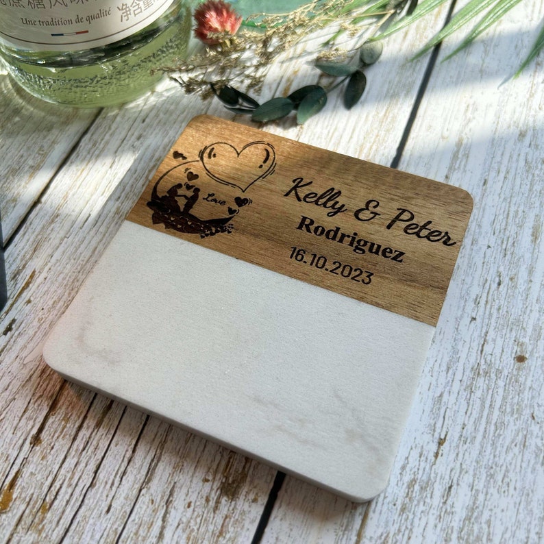 Pu&ograve; includere: Un sottobicchiere bianco e in legno con un design a forma di cuore e il testo "Kelly & Peter Rodriguez 16.10.2023" inciso sul legno.