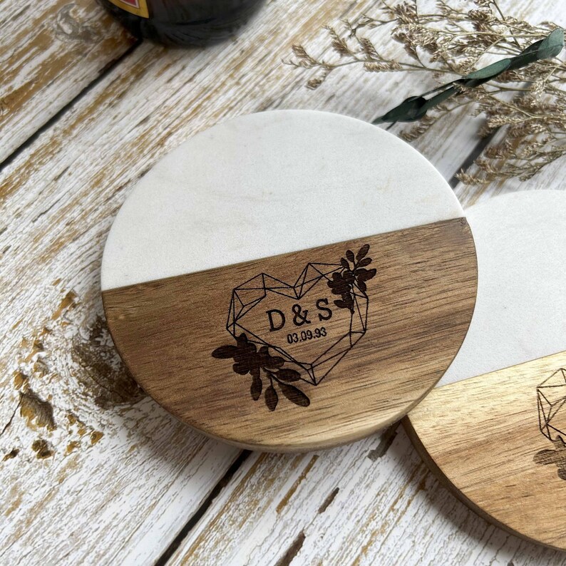 Pu&ograve; includere: Sottobicchiere rotondo in legno e marmo con un design a cuore geometrico e le iniziali "D&S" e la data "03.09.93" incise sul legno.