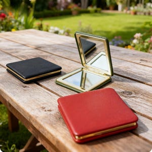 May include: Three square compact mirrors in black, red, and green leather with gold-colored trim. One mirror is open, revealing a reflective surface. The mirrors are arranged on a wooden table outdoors, with a blurred background of greenery.