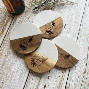 Pu&ograve; includere: Quattro sottobicchieri rotondi con un top in marmo bianco e un fondo in legno. Ogni sottobicchiere presenta un design a forma di cuore con iniziali e una data incise sul fondo in legno. Le iniziali sono D&S e D&M.