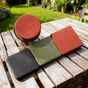 May include: A collection of coasters in various shapes and colours. There is a stack of round, red coasters and three square coasters in black, green, and red. They are on a wooden table, with a garden in the background.
