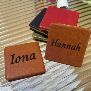 Pode incluir: Uma coleção de espelhos compactos quadrados com capas texturizadas macias em várias cores, incluindo castanho claro, preto, vermelho e verde azeitona. Dois espelhos castanhos estão em destaque, com os nomes Iona e Hannah gravados.