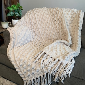 May include: A cream-colored crocheted throw blanket with a textured pattern and fringe. The blanket is draped over a couch.