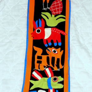 May include: A colorful textile wall hanging with a black background features a variety of animals and a pineapple. The animals include a bird, a donkey, a dog, and a fish. The textile is framed with a blue and orange border.