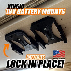 Może przedstawiać: Dwa czarne plastikowe uchwyty na baterie do baterii Ridgid 18V. Uchwyty są przymocowane do drewnianej powierzchni i mają nadrukowany tekst "RIDGID". Tekst "BATTERIES LOCK IN PLACE!" jest nadrukowany pod uchwytami, wraz z małą flagą amerykańską i tekstem "Made in USA".