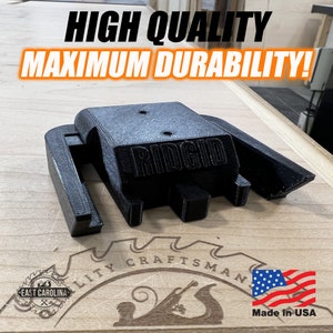 May include: A black 3D printed Ridgid tool holder, made in the USA. The holder is designed to fit a Ridgid tool and has a slot for a belt clip. The holder is sitting on a wooden surface with a Craftsman logo and a US flag.