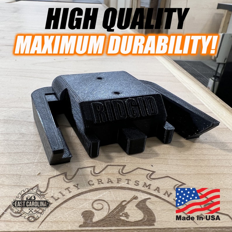 May include: A black 3D printed Ridgid tool holder, made in the USA. The holder is designed to fit a Ridgid tool and has a slot for a belt clip. The holder is sitting on a wooden surface with a Craftsman logo and a US flag.