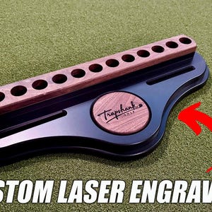 May include: A black golf ball holder with a wooden top featuring multiple holes. The holder has a circular wooden accent with the "Traphank Golf" logo. The text "CUSTOM LASER ENGRAVING" is at the bottom.