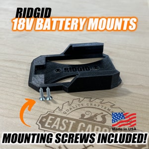 Może przedstawiać: Czarny plastikowy uchwyt do baterii 18V do narzędzi RIDGID. Uchwyt zawiera dwa śruby montażowe. "RIDGID" jest nadrukowane na uchwycie. "Made in USA" jest nadrukowane na naklejce.