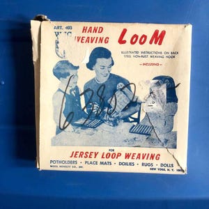 Peut inclure: Une boîte en carton vintage pour un métier à tisser manuel, Art. 403, par Wool Novelty Co., Inc. L'emballage crème présente des illustrations bleues d'une personne et de deux enfants tissant, avec du texte rouge indiquant Hand Weaving Loom for Jersey Loop Weaving.