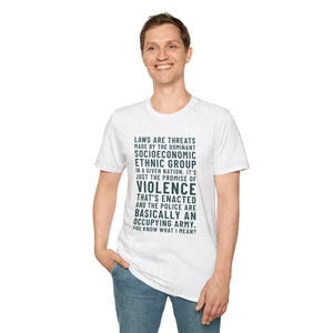 May include: White t-shirt with a crew neck featuring a printed message in dark teal. The text reads: "LAWS ARE THREATS MADE BY THE DOMINANT SOCIOECONOMIC ETHNIC GROUP...YOU KNOW WHAT I MEAN?" The shirt is worn by a person.