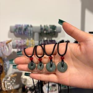 Könnte beinhalten: Vier Jade-Donut-Anhänger an schwarzen Kordeln, jeweils mit einem roten Perlenakzent. Die Anhänger variieren leicht in der Größe und werden in einer Hand gehalten. Im Hintergrund sind weitere Schmuckstücke zu sehen.