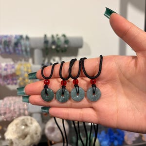 May include: Four jade pendant necklaces with black cords and red beads. Each pendant is a circular, light green stone with a hole in the center. The necklaces are displayed on a hand, with other jewelry visible in the background.