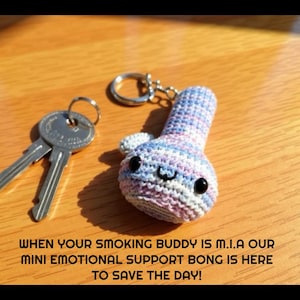 May include: A handmade crochet bong keychain in blue, pink, and white, with a smiling face and black eyes. The keychain is next to a set of keys on a wooden surface. Text on the image reads: "WHEN YOUR SMOKING BUDDY IS M.I.A OUR MINI EMOTIONAL SUPPORT BONG IS HERE TO SAVE THE DAY!"