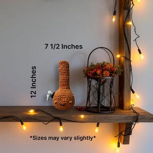 May include: A crocheted orange bong, approximately 12 inches tall and 7.5 inches wide, with a white mouthpiece. The text on the image reads "Sizes may vary slightly". The bong is displayed on a wooden shelf with a decorative metal basket filled with flowers.