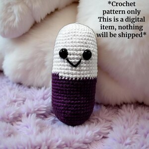 May include: A crocheted pill-shaped plush toy with a white top and purple bottom. The toy has black eyes and a smiling mouth.