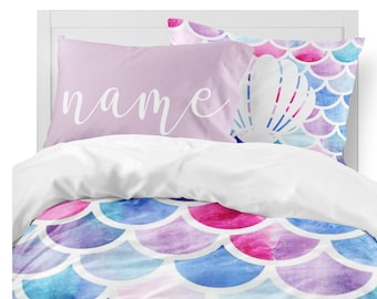 Könnte beinhalten: Ein lila-weißes Bettwäsche-Set mit Meerjungfrauen-Motiv und einem Kissen mit der Aufschrift "name". Die Bettwäsche zeigt ein Aquarell-Design mit Meerjungfrauen-Schuppen und einer Muschel.